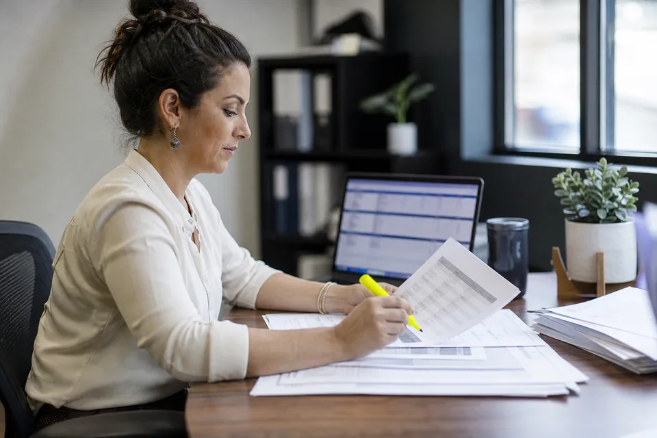 Underwriter at a merchant cash advance funder reviewing printed business bank statements beside a laptop